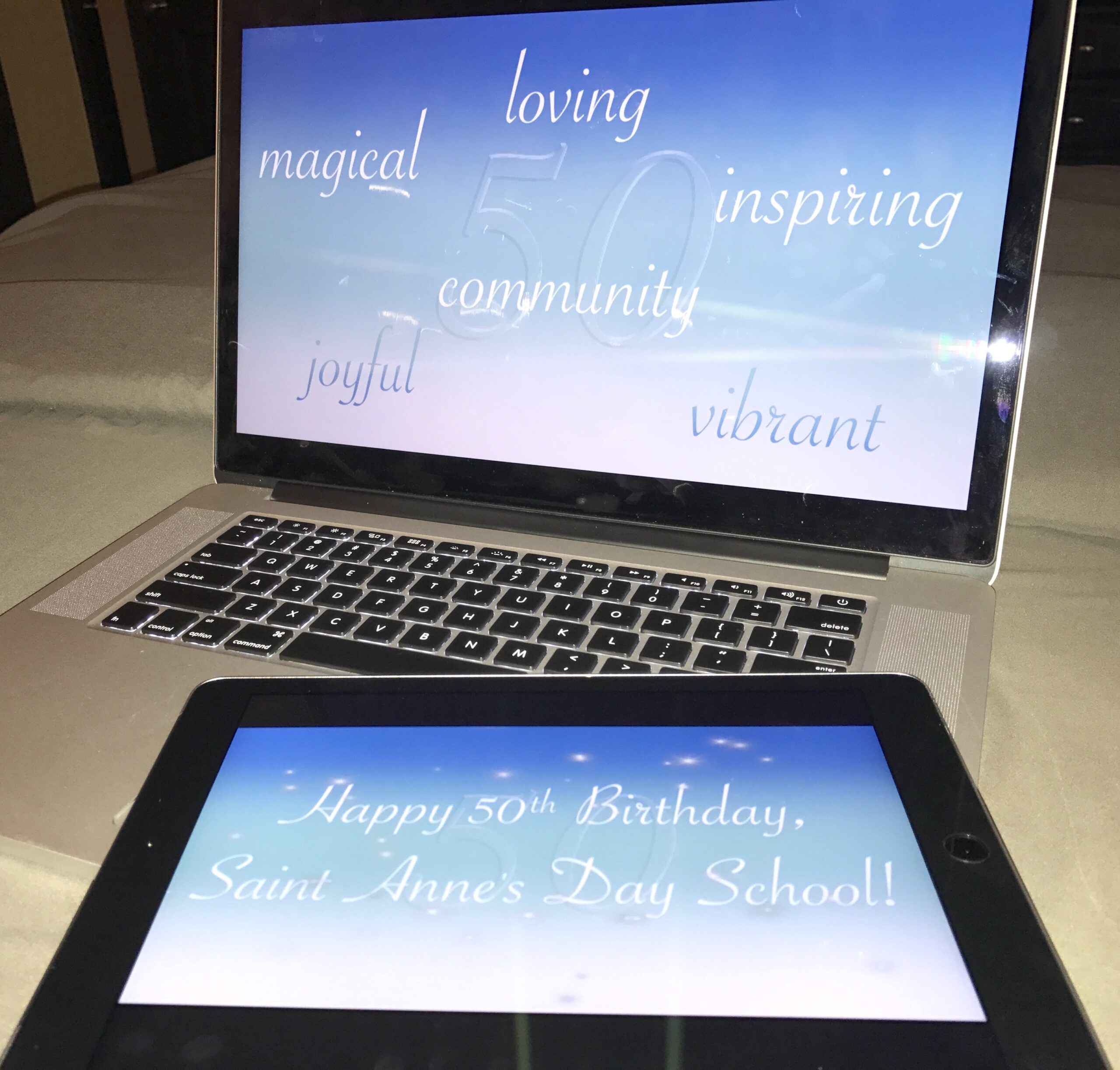 open laptop computer with large iPad laying angled on the keyboard, with both screens displaying the opening title card of Saint Anne's 50th Birthday video